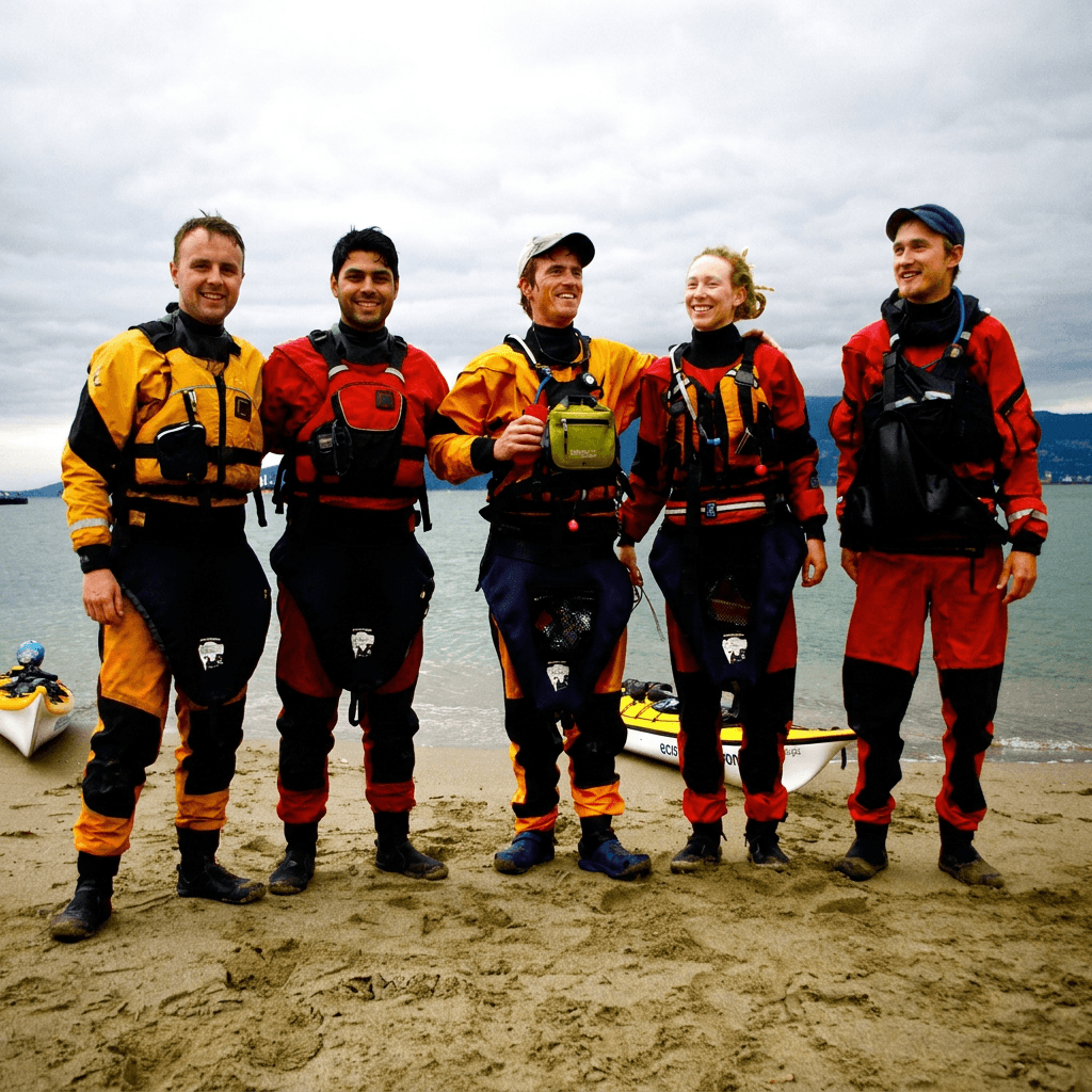 The kayak expedition team on the beach, drysuits on, kayaks behind them